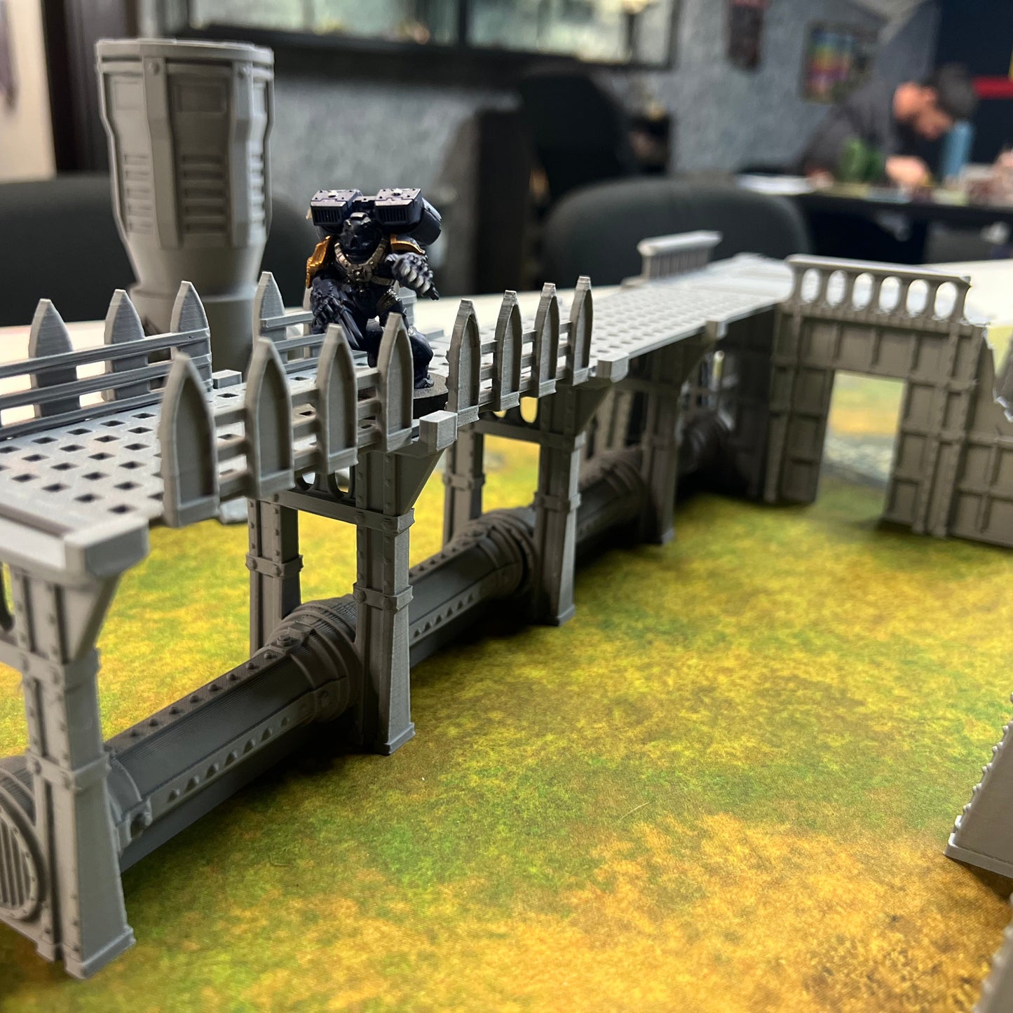 A gray plastic miniature bridge or walkway structure displayed on a green and yellow tabletop terrain mat, with a small black and blue toy figure standing on top. The background shows a blurred indoor office or workshop setting with chairs and a desk.