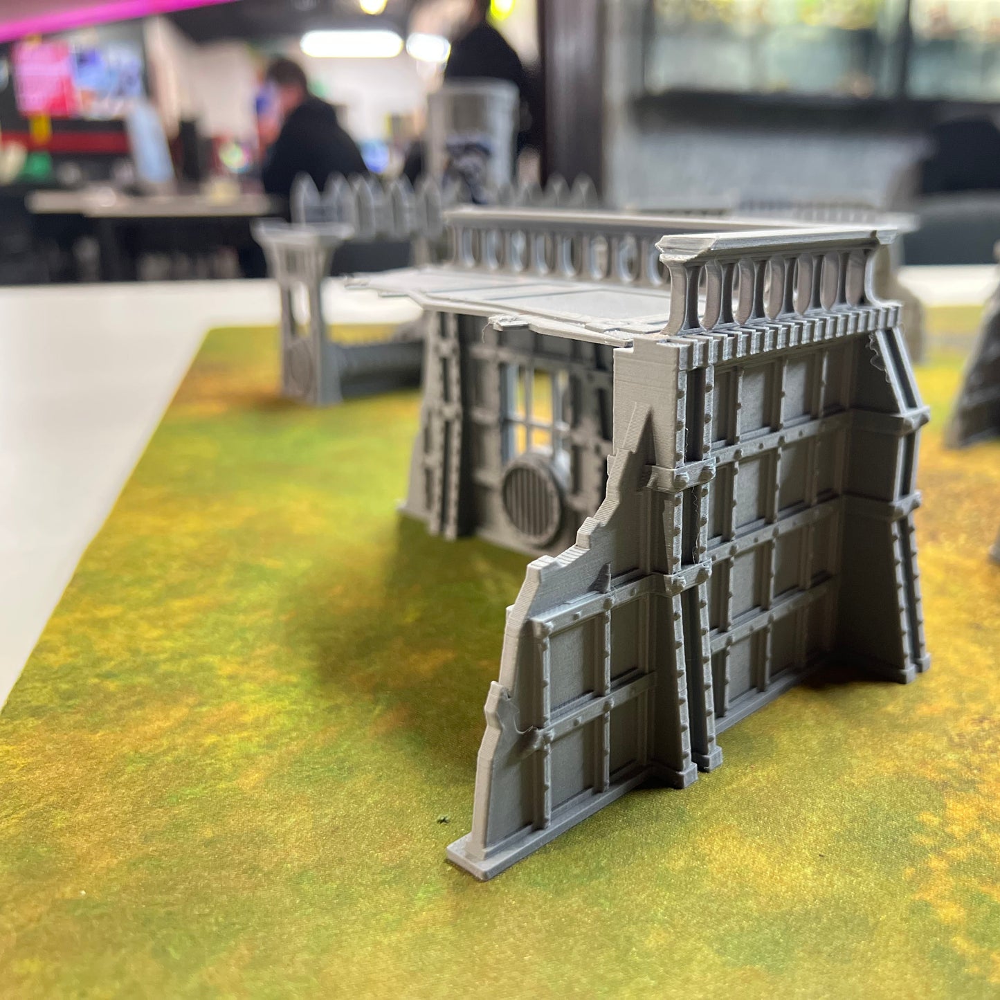 Gray plastic miniature terrain wall pieces displayed on a green and yellow tabletop gaming mat, with a blurred indoor tabletop setup in the background.