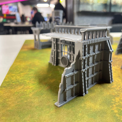 Gray plastic miniature terrain wall pieces displayed on a green and yellow tabletop gaming mat, with a blurred indoor tabletop setup in the background.