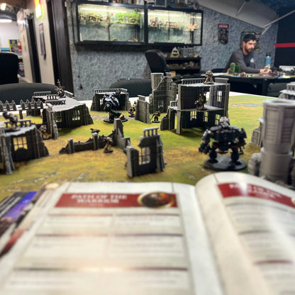 A tabletop gaming scene with miniature terrain pieces and structures arranged on a green battle mat, with an open rulebook in the foreground and people seated at tables in the background inside what appears to be a hobby shop or gaming lounge.