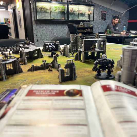 A tabletop gaming scene with miniature terrain pieces and structures arranged on a green battle mat, with an open rulebook in the foreground and people seated at tables in the background inside what appears to be a hobby shop or gaming lounge.