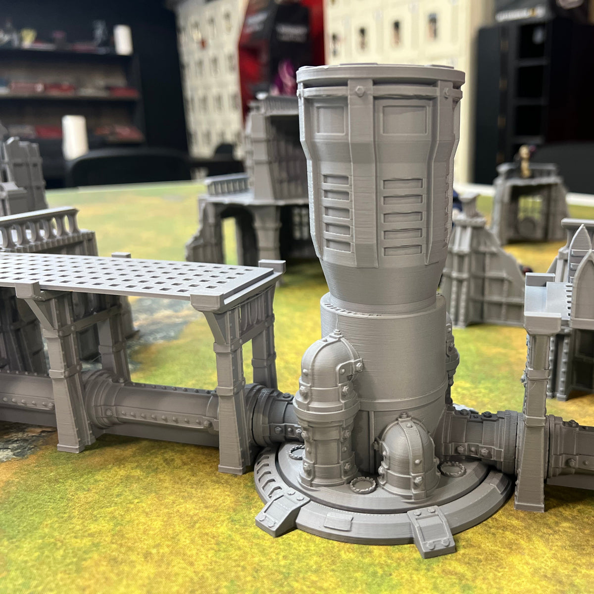 A gray unpainted miniature building structure, likely a tower or industrial building component, displayed on a green and yellow tabletop gaming mat with other miniature buildings in the background.