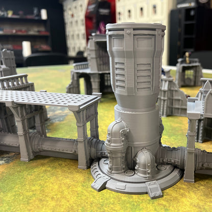 A gray unpainted miniature building structure, likely a tower or industrial building component, displayed on a green and yellow tabletop gaming mat with other miniature buildings in the background.